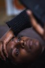 Зверху серйозно продумана молода етнічна жінка в повсякденному теплому светрі розслабляється на зручному м'якому дивані і посміхається при використанні смартфона — стокове фото