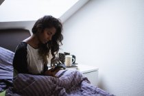 Vue latérale de femme indienne réfléchie en vêtements de nuit assis sur un lit confortable sur un oreiller doux et une couverture en utilisant le téléphone portable dans la chambre à coucher lumineuse — Photo de stock