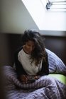 Vista lateral de la mujer india reflexiva en ropa de dormir sentado en la cama acogedora en almohada suave y manta usando el teléfono móvil en el dormitorio ligero - foto de stock