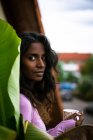 Красивая этническая женщина с длинными темными волосами в розовой повседневной одежде, держа чашку горячего напитка в руках, стоя на балконе возле зеленого листа растения, глядя в камеру — стоковое фото