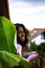 Позитивна молода етнічна жінка з довгим темним волоссям в рожевому повсякденному одязі тримає чашку свіжого гарячого напою в руках, стоячи на балконі біля рослини зеленого листа, дивлячись на камеру — стокове фото