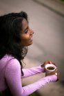 De cima vista da jovem senhora étnica positiva com cabelo escuro longo vestindo roupas casuais rosa segurando xícara de bebida quente fresca nas mãos enquanto estava na varanda e olhando para longe — Fotografia de Stock