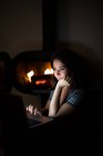 Donna pensierosa in maglietta casual seduta in camera buia accogliente vicino al camino e leggere libro su netbook mentre riposa durante la notte — Foto stock