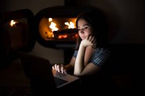 Mulher pensativa em camiseta casual sentada no quarto escuro aconchegante perto da lareira e lendo livro no netbook enquanto descansa durante a noite — Fotografia de Stock