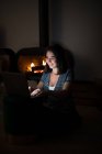 Pensive female in casual t shirt sitting in dark cozy room near fireplace and reading book on netbook while resting during night time — Stock Photo