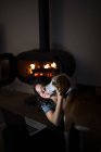 Зверху щаслива жінка сидить на підлозі у темній вітальні з каміном і обіймає Хар'єра, дивлячись відео на ноутбук разом — стокове фото