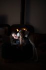 Сверху счастливая женщина сидит на полу в темной гостиной с камином и обнимает Харриера, смотря видео на ноутбуке вместе — стоковое фото