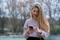 Расслабленная молодая женщина в случайном джампере смотрит социальные сети на смартфоне и улыбается, сидя на каменной скамейке на улице — стоковое фото