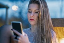 Giovane ragazza che usa il cellulare di notte appoggiata a una panchina — Foto stock