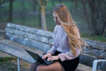 Mujer joven en ropa casual y con el pelo que fluye sentado en el banco en el parque y responder a correo electrónico mientras escribe en el teclado del ordenador portátil - foto de stock