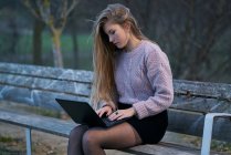 Молодая женщина в повседневной одежде и с текущими волосами, сидя на скамейке в парке и отвечая на электронную почту во время ввода на клавиатуре ноутбука — стоковое фото