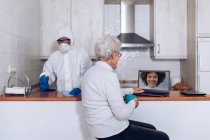 Ältere Frau plaudert auf Laptop und Hausangestellte desinfiziert Küche während Pandemie — Stockfoto