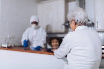 Femme âgée bavardant sur un ordinateur portable et un employé des soins à domicile désinfectant la cuisine pendant une pandémie — Photo de stock