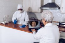 Пожилая женщина болтает на ноутбуке и на дому работник дезинфекции кухни во время пандемии — стоковое фото