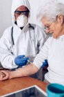 Professional male caregiver in protective uniform and gloves visiting aged female client at home and making injection during coronavirus outbreak — Stock Photo
