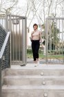 Erwachsene glückliche brünette Frau in lässigem Outfit und grauem Schal tritt an sonnigen Tagen auf Betonpflastertreppen und schaut weg — Stockfoto
