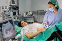 Doctora en máscara estéril y guante azul usando escáner de ultrasonido mientras examina a una mujer embarazada alegre y mira la pantalla de la computadora en el hospital - foto de stock