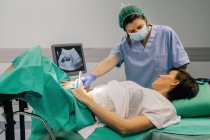 Doctora en máscara estéril y guante azul usando escáner de ultrasonido mientras examina a una mujer embarazada alegre en el hospital - foto de stock