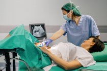 Женщина-врач в стерильной маске и голубой перчатке с помощью ультразвукового сканера при обследовании веселой беременной женщины и при взгляде на экран компьютера в больнице — стоковое фото