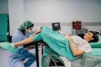Vista laterale del medico donna in uniforme blu e maschera sterile esaminando paziente sorridente sulla sedia ginecologica in clinica di fertilità — Foto stock