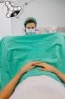 Жінка-лікар у блакитній формі та стерильній масці, яка вивчає невпізнаваного пацієнта на гінекологічному кріслі в клініці фертильності — стокове фото