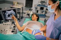 Doctora en máscara estéril y guante azul usando escáner de ultrasonido mientras examina a una mujer embarazada alegre apuntando a la pantalla en el hospital - foto de stock