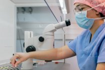 Вид сбоку женщины в медицинской форме и маске с помощью современного микроскопа для исследования клеток человека во время работы в лаборатории современной клиники — стоковое фото