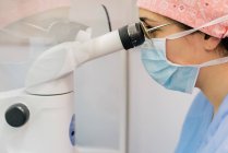 Visão lateral da mulher de uniforme médico e máscara usando microscópio moderno para examinar células humanas enquanto trabalhava no laboratório da clínica moderna — Fotografia de Stock