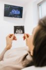 Mujer embarazada irreconocible que inspecciona la imagen de la ecografía mientras está acostada en la cama en la sala de la clínica moderna de fertilidad - foto de stock