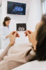 Femme enceinte méconnaissable inspectant l'image d'échographie alors qu'elle était allongée sur le lit dans la salle de la clinique de fertilité moderne — Photo de stock