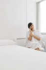 Веселая женщина в белом халате сидит на кровати возле кровати и пользуется смартфоном в подопечной современной клиники — стоковое фото