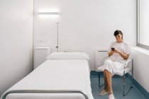 Mujer alegre en bata blanca sentada en la cama cerca de la cama y el teléfono inteligente de navegación en la sala de la clínica moderna - foto de stock