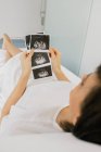Зверху вагітна жінка оглядає фотографію сонограми, лежачи на ліжку в палаті сучасної клініки фертильності — стокове фото