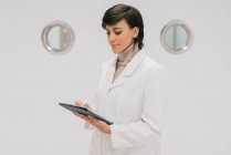 Visão lateral do médico positivo usando tablet enquanto está de pé contra a porta branca no corredor do hospital moderno — Fotografia de Stock