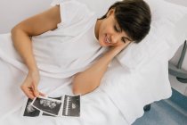 De cima grávida do sexo feminino inspecionando imagem de ultrassonografia enquanto deitado na cama na ala da clínica de fertilidade moderna — Fotografia de Stock