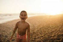 Joyeux garçon torse nu souriant et regardant la caméra tout en se tenant sur la plage de sable pendant le coucher du soleil — Photo de stock