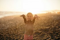 Щасливий без сорочки хлопчик посміхається і дивиться на камеру, стоячи на піщаному пляжі під час заходу сонця — стокове фото