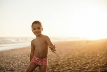 Happy shirtless boy smiling and looking at camera while standing on sandy beach during sunset — Stock Photo