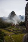 Increíble vista del verde valle rocoso con vallas de piedra y pequeñas casas de piedra ubicadas cerca del pico cubierto por nubes de niebla en Perú - foto de stock