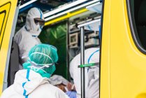 Вид сбоку анонимных врачей в защитном костюме, стоящих в машине скорой помощи рядом с открытой дверью с оборудованием и осматривающих пациента с вирусной инфекцией — стоковое фото