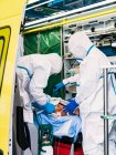 Вид збоку анонімних лікарів у захисному костюмі, що стоїть на машині швидкої допомоги біля відкритих дверей з обладнанням та вивчає пацієнта з вірусною інфекцією — стокове фото