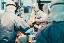 Médicos profissionais irreconhecíveis em uniformes e máscaras protetoras cuidando de pacientes com infecção viral em pé na sala de cirurgia no hospital moderno — Fotografia de Stock