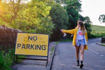 Щаслива жінка в жовтій куртці і джинсових шортах показує дорожній знак Без паркування під час проходження по сільській дорозі в сонячний літній день — стокове фото