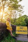 Mujer en chaqueta amarilla y pantalones cortos de mezclilla apoyada en una pared limpia una señal de tráfico No hay aparcamiento en la carretera rural en la tarde soleada de verano - foto de stock