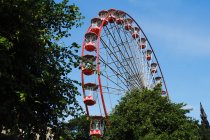 Из-под колеса обозрения с красными каютами, расположенными в парке развлечений с деревьями и башней в солнечный день с голубым небом — стоковое фото