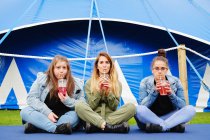 Cheerful young women in casual clothes drinking red beverage through straw while sitting on blue mat near fenced tent — Stock Photo