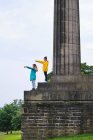 Вид збоку на жінок у різнокольорових куртках, що стоять на сходинках пам'ятника та вказують пальцем у небі на сонячний день у парку — стокове фото
