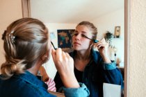 Vista posterior de la mujer enfocada en chaqueta de mezclilla mirando en el espejo y la aplicación de rimel maquillaje contra el interior borroso de acogedor apartamento moderno - foto de stock