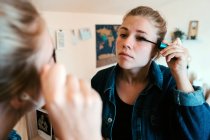 Back view of focused female in denim jacket looking in mirror and applying mascara make up against blurred interior of cozy modern apartment — Stock Photo