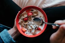 From above crop female in casual clothes holding metal spoon and red bowl with pieces of strawberry and oatmeal poured with milk while sitting on bed in light bedroom — Stock Photo